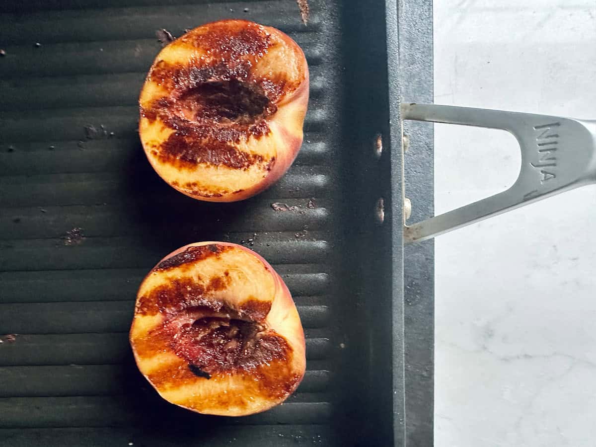 Two grilled peach halves with visible char marks are on a black ridged grill pan, next to a metal handle labeled Ninja, set against a light-colored countertop.