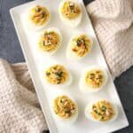 A rectangular white plate with eight Honey-Mustard Deviled Eggs, garnished with herbs and crumbled topping, sits on a gray surface with beige textured towels and a bowl of breadsticks nearby.