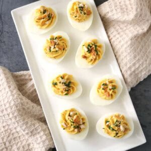 A rectangular white plate with eight Honey-Mustard Deviled Eggs, garnished with herbs and crumbled topping, sits on a gray surface with beige textured towels and a bowl of breadsticks nearby.