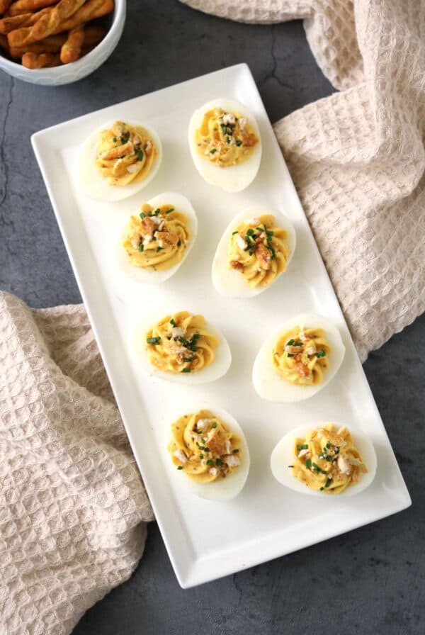 A rectangular white plate with eight Honey-Mustard Deviled Eggs, garnished with herbs and crumbled topping, sits on a gray surface with beige textured towels and a bowl of breadsticks nearby.