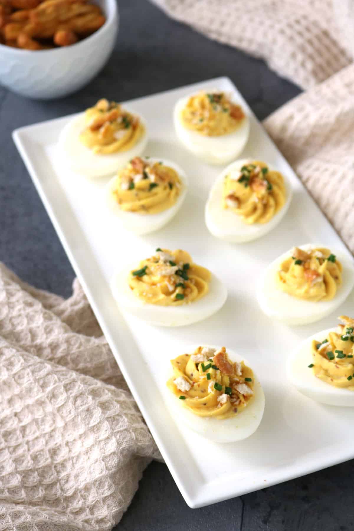 Eight Honey-Mustard Deviled Eggs, topped with herbs and crispy bits, are arranged on a white rectangular plate. A beige textured cloth and a bowl of snacks are visible in the background.