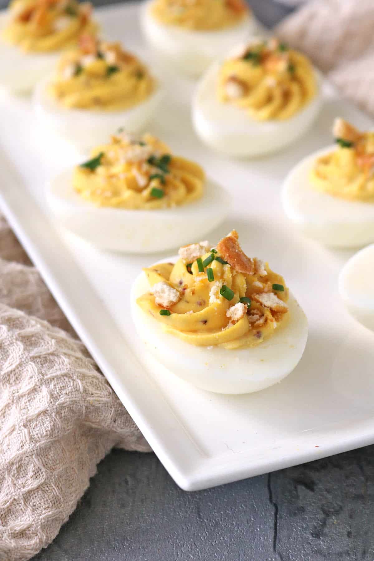 A white platter with Honey-Mustard Deviled Eggs, topped with chopped chives and crushed crackers, set on a gray surface next to a beige textured cloth.