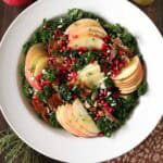 A white bowl filled with kale salad topped with apple slices, pomegranate seeds, nuts, prosciutto, and goat cheese. A glass jar of dressing, pomegranate, apples, and herbs are nearby on a wooden table.
