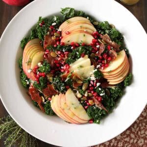A white bowl filled with kale salad topped with apple slices, pomegranate seeds, nuts, prosciutto, and goat cheese. A glass jar of dressing, pomegranate, apples, and herbs are nearby on a wooden table.