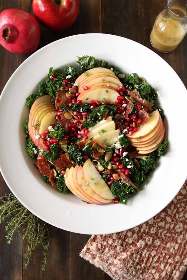 A white bowl filled with kale salad topped with apple slices, pomegranate seeds, nuts, prosciutto, and goat cheese. A glass jar of dressing, pomegranate, apples, and herbs are nearby on a wooden table.
