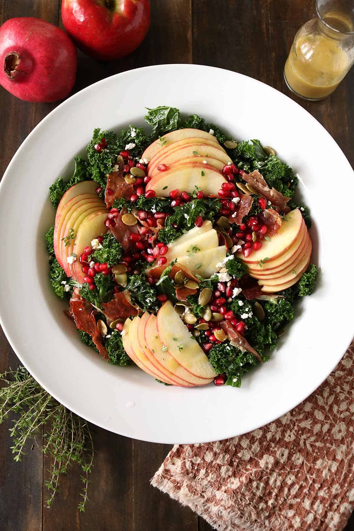A white bowl filled with kale salad topped with apple slices, pomegranate seeds, nuts, prosciutto, and goat cheese. A glass jar of dressing, pomegranate, apples, and herbs are nearby on a wooden table.