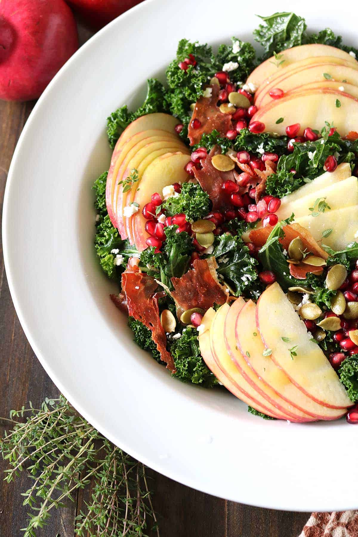 A salad with kale, thin apple slices, pomegranate seeds, pumpkin seeds, crispy prosciutto, and fresh thyme, served in a white bowl on a wooden table.