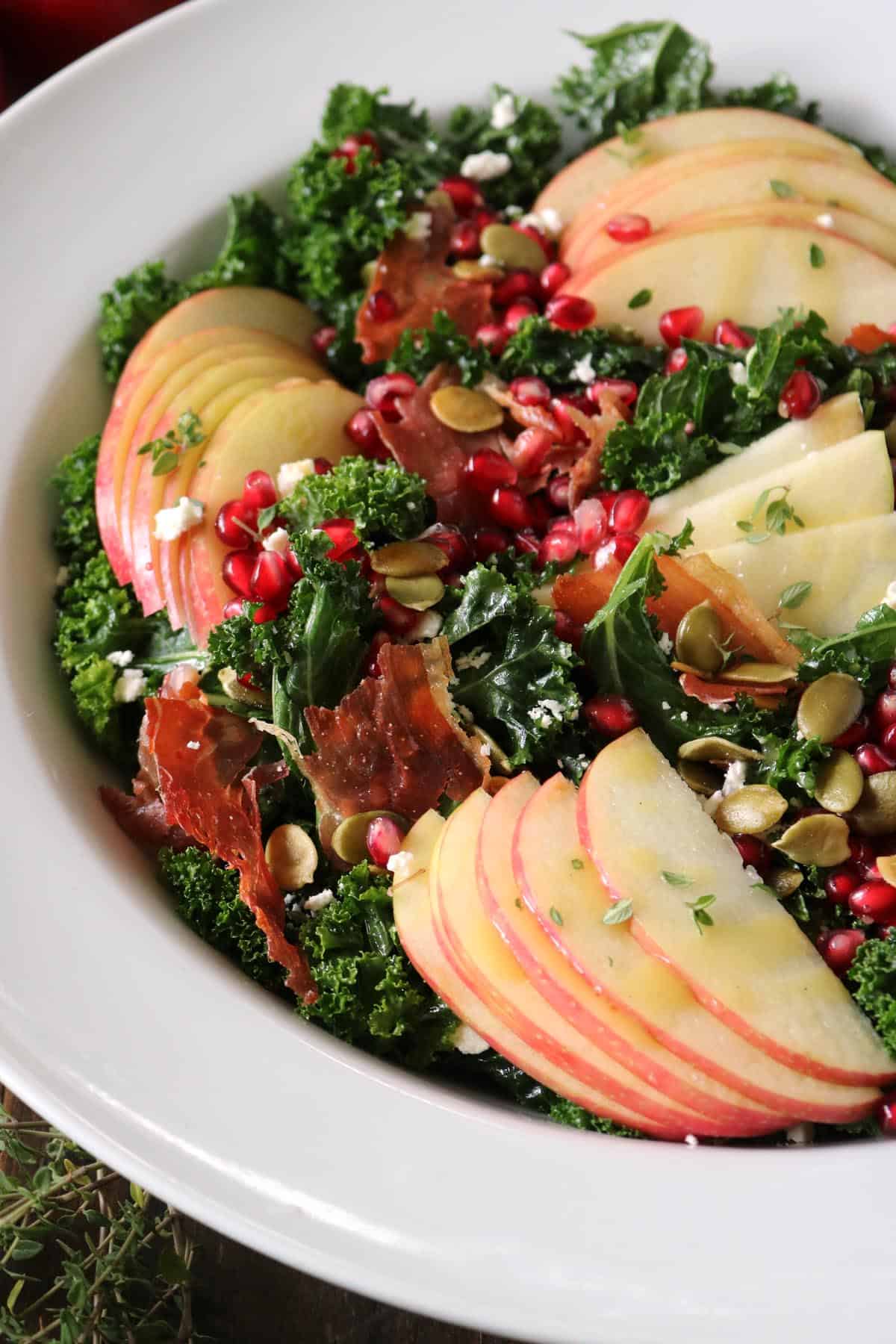 A salad with kale, apple slices, pomegranate seeds, pumpkin seeds, prosciutto, and herbs, served in a white bowl.