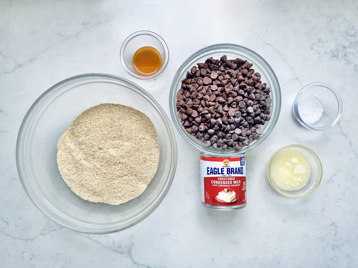 Ingredients for 1-2-3 Snap Brownies displayed on a white countertop, including a bowl of graham cracker crumbs, a bowl of chocolate chips, can of sweetened condensed milk, melted butter, vanilla extract, and a bowl of salt.