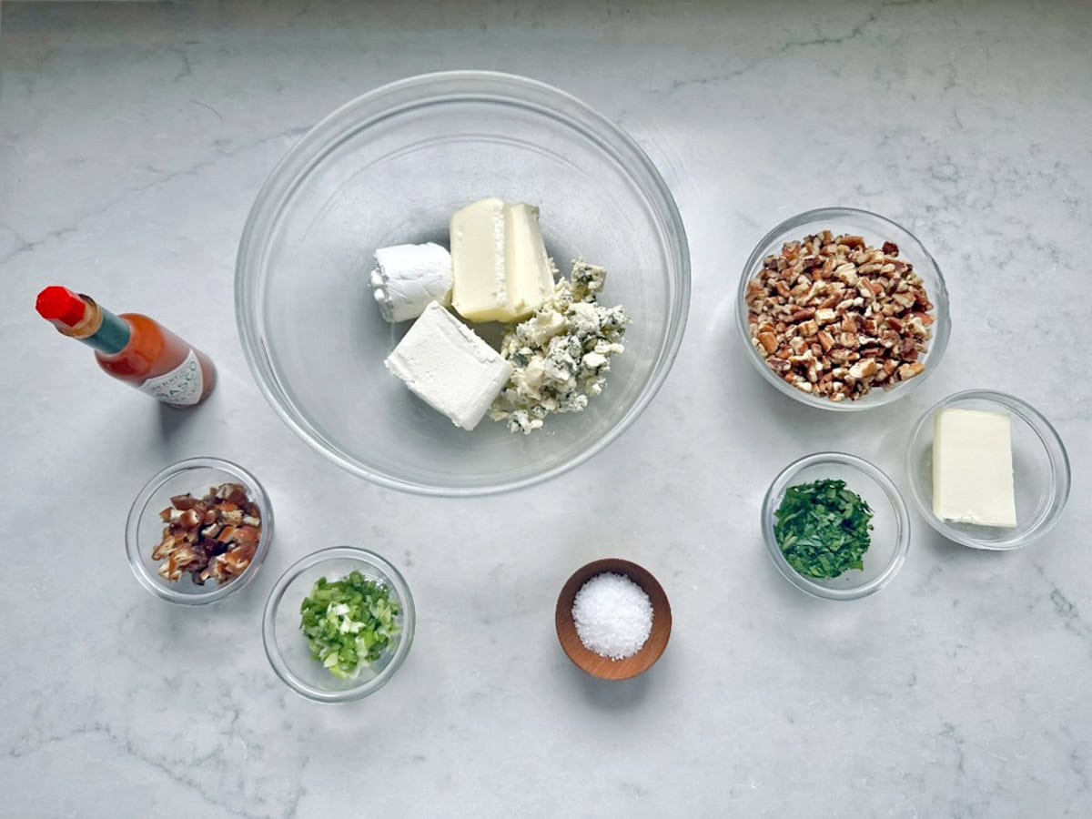 A glass bowl with cheeses and butter, inspired by Vivian Howard’s Party Magnet, is surrounded by small bowls of chopped nuts, herbs, dates, green onions, salt, and a bottle of hot sauce on a marble surface.