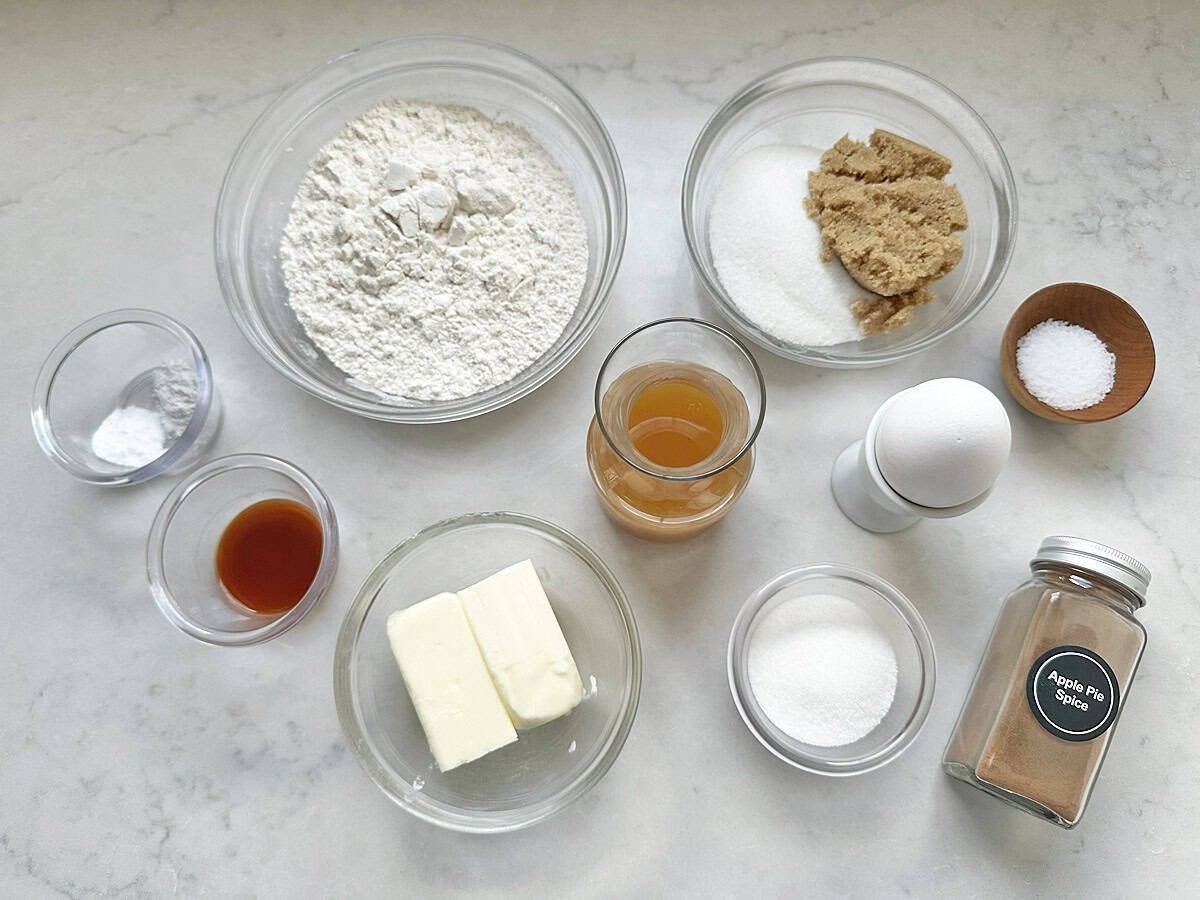 Baking ingredients on a counter: flour, brown and white sugars, salt, egg, butter sticks, vanilla extract, apple cider, baking soda, and a jar labeled Apple Pie Spice—everything you need to whip up delicious Apple Cider Snickerdoodles.