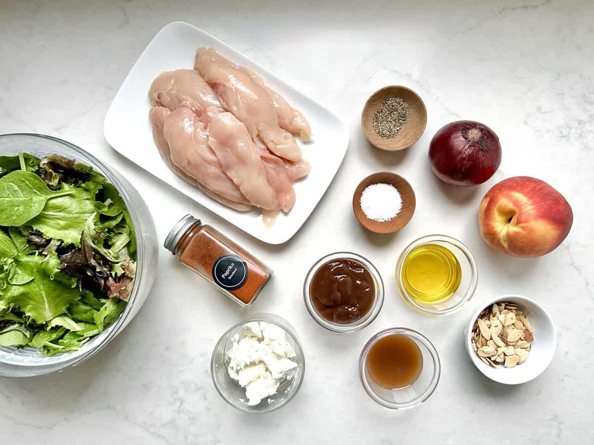 Fresh salad greens, raw chicken breasts on a white plate, a red onion, apple, spices, paprika, feta cheese, vinaigrette, BBQ sauce, olive oil, and sliced almonds arranged on a white countertop.