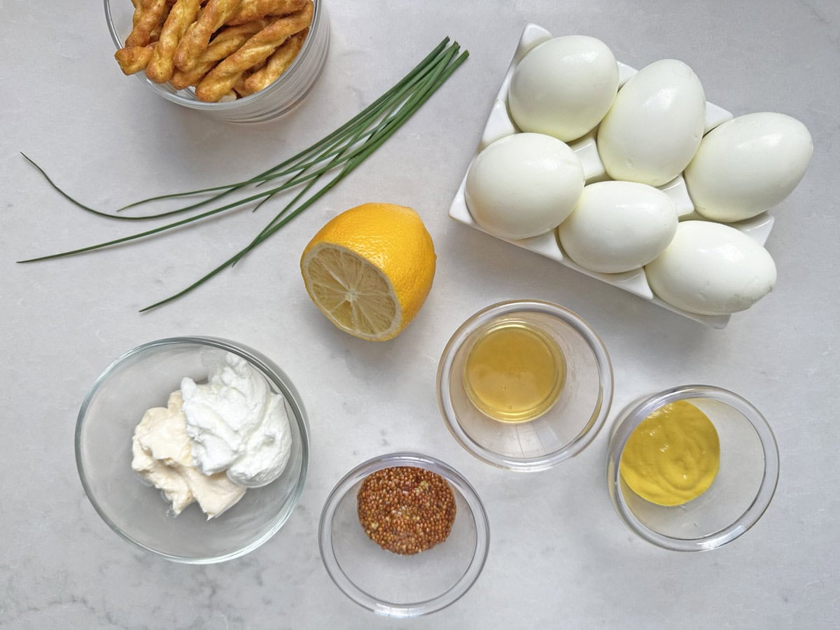 Ingredients for Honey-Mustard Deviled Eggs: hard-boiled eggs, chives, lemon half, breadsticks, mayonnaise, yogurt, whole grain mustard, Dijon mustard, and apple cider vinegar on a white surface.