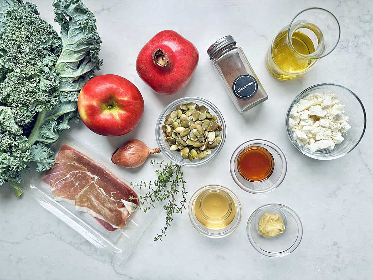 Various fresh ingredients are arranged on a white surface, ready for a delicious Kale Apple Harvest Salad. The selection includes kale, an apple, a pomegranate, prosciutto, feta cheese, seasonings, seeds, a shallot, olive oil, and small bowls of liquids and mustard.