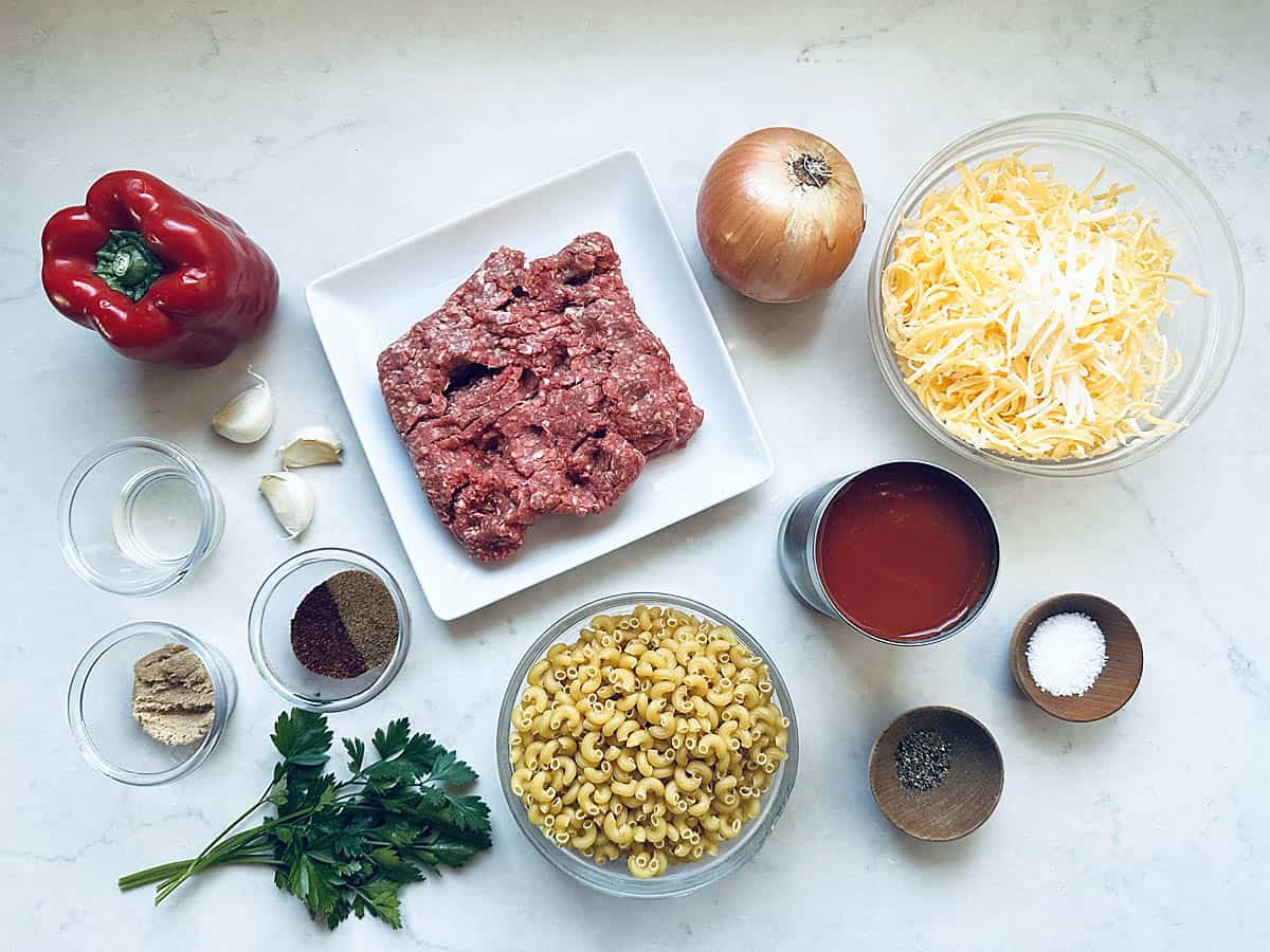 Flat lay of Skillet Cheesy Chili Mac ingredients: ground beef, elbow macaroni, shredded cheese, tomato sauce, onion, red bell pepper, garlic, parsley, spices, salt, pepper, and seasonings on a white countertop.