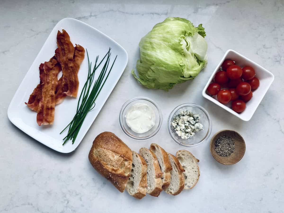 Sliced bread, cooked bacon, chives, iceberg lettuce, cherry tomatoes, blue cheese crumbles, mayonnaise, and cracked black pepper arranged neatly on a white surface.