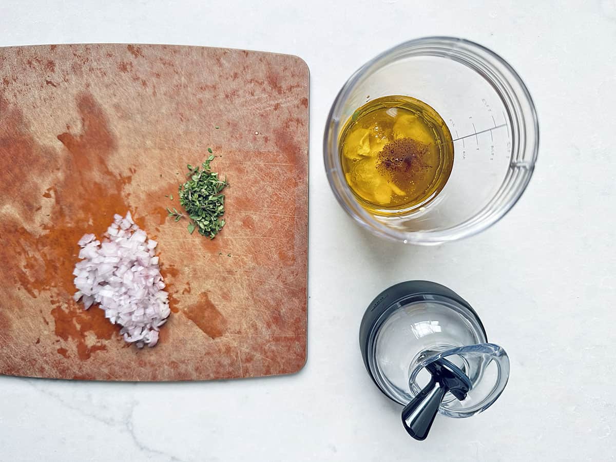 Chopped shallots and herbs on a wooden cutting board next to a blender cup filled with oil, vinegar, and seasoning—perfect for preparing the dressing that will elevate your Kale Apple Harvest Salad—sit alongside a blender lid on a white surface.