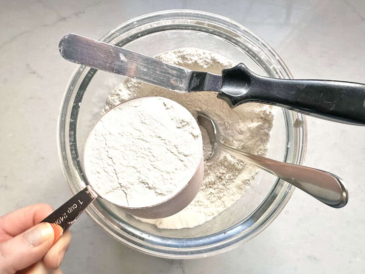 A hand levels a 1-cup measuring cup of flour with a knife over a glass bowl filled with flour, prepping the ingredients for Apple Cider Snickerdoodles. A spoon rests inside the bowl on a light countertop.