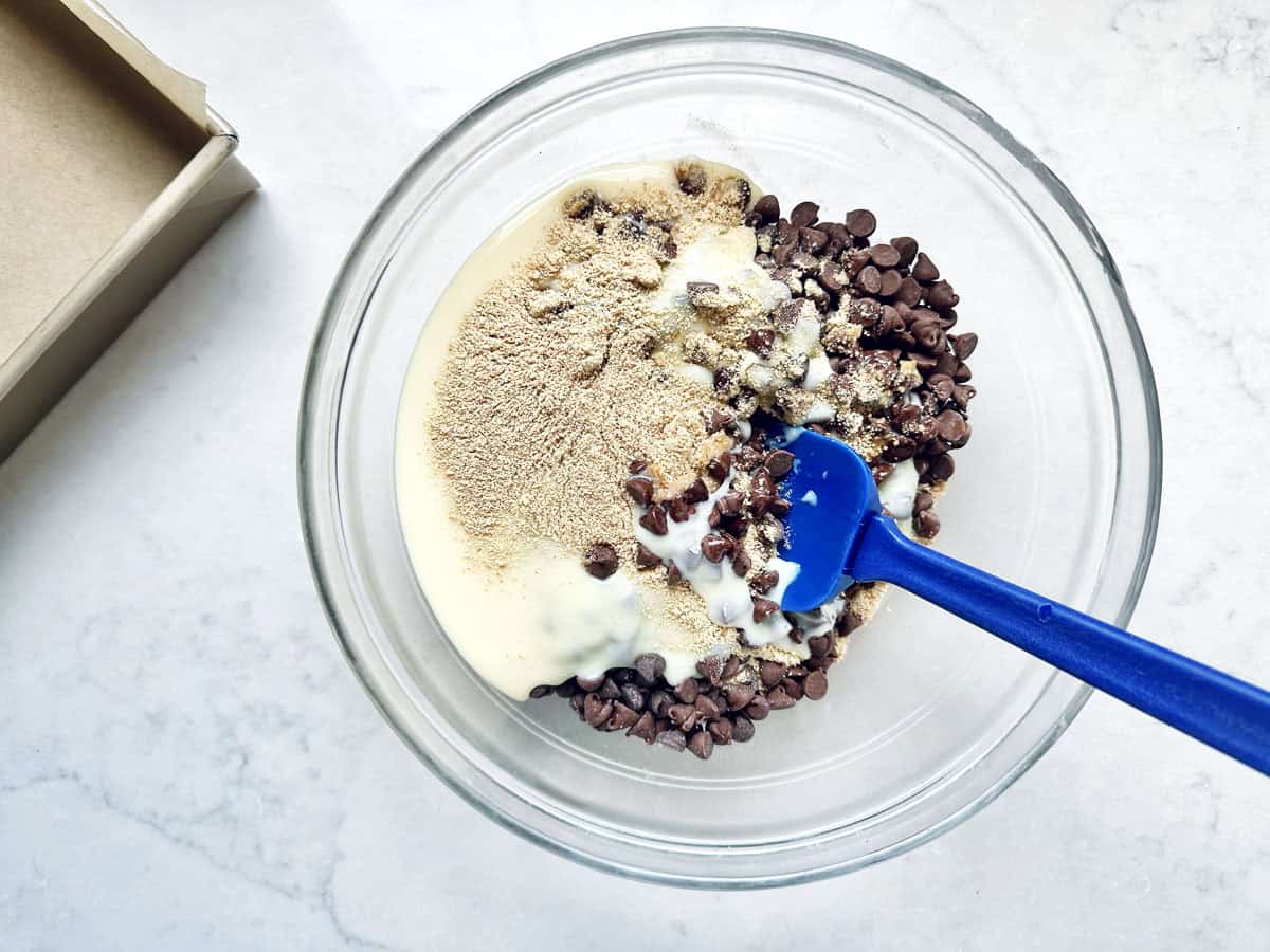 Top-down view of a clear glass bowl containing the ingredients for 1-2-3 Snap Brownies: chocolate chips, condensed milk, and graham cracker crumbs. A blue rubber spatula is inside the bowl, and a lined baking pan sits in the upper-left corner on a white marble surface.