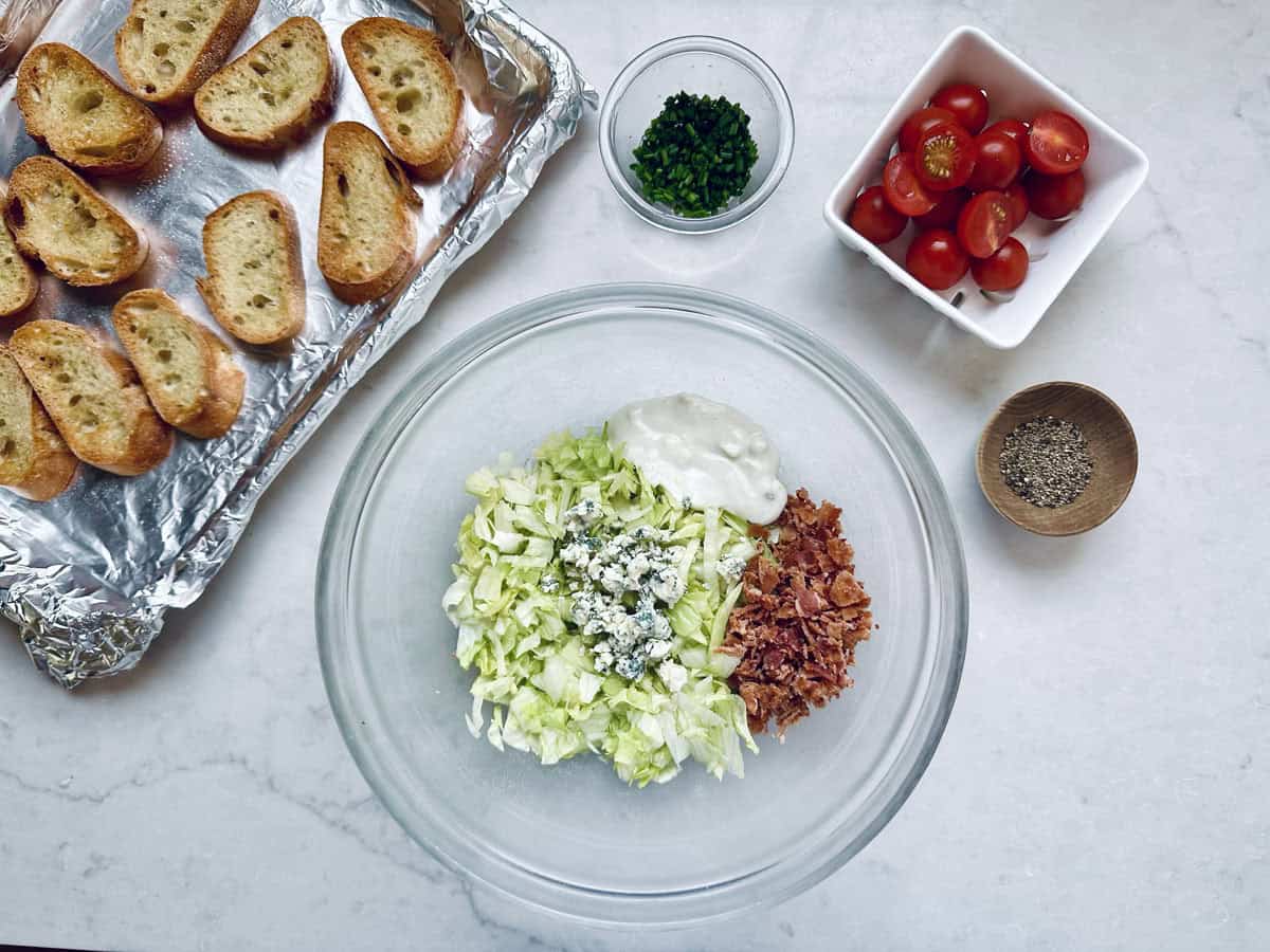 A glass bowl with chopped lettuce, bacon bits, blue cheese, and dressing on a white surface, surrounded by toasted bread, cherry tomatoes, chopped herbs, and a small bowl of black pepper.