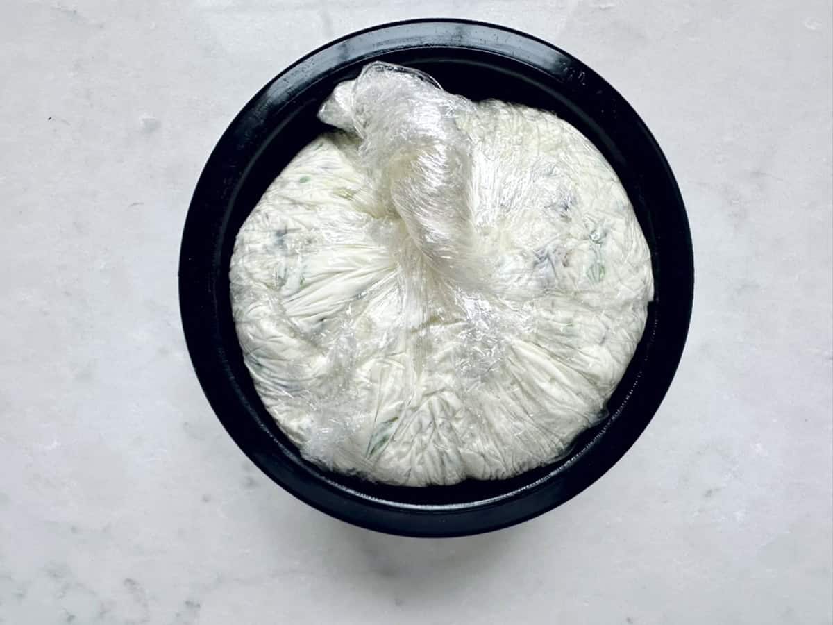A round black bowl contains dough wrapped tightly in plastic wrap, resting on a light-colored surface—just the kind of prep you might find before making Vivian Howard’s Party Magnet.