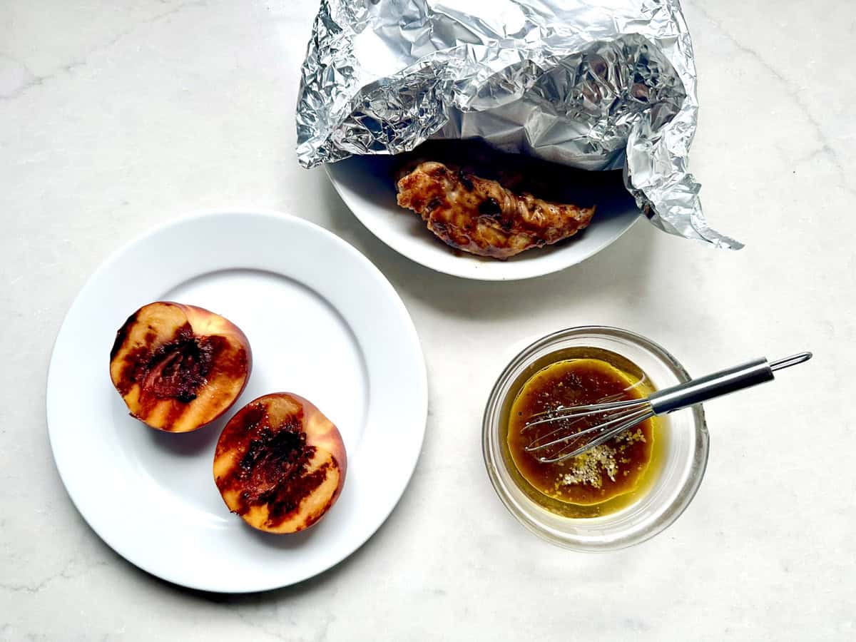 A grilled peach cut in half on a white plate, a piece of cooked meat partially wrapped in foil on another plate, and a small bowl of sauce with a whisk on a light surface.
