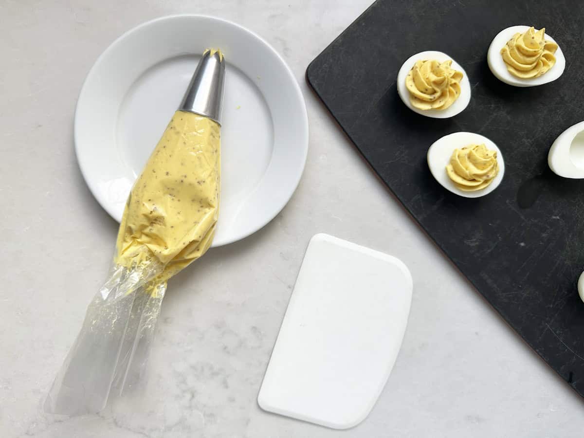 A piping bag filled with yellow Honey-Mustard Deviled Egg filling rests on a white plate beside a black tray of halved eggs with piped yolk mixture. A white scraper tool lies nearby on the countertop.