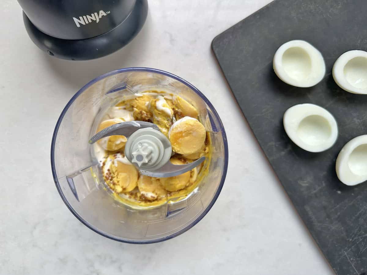 A food processor with egg yolks, mustard, and mayo inside sits next to a black cutting board holding empty egg white halves—perfect for assembling delicious Honey-Mustard Deviled Eggs.