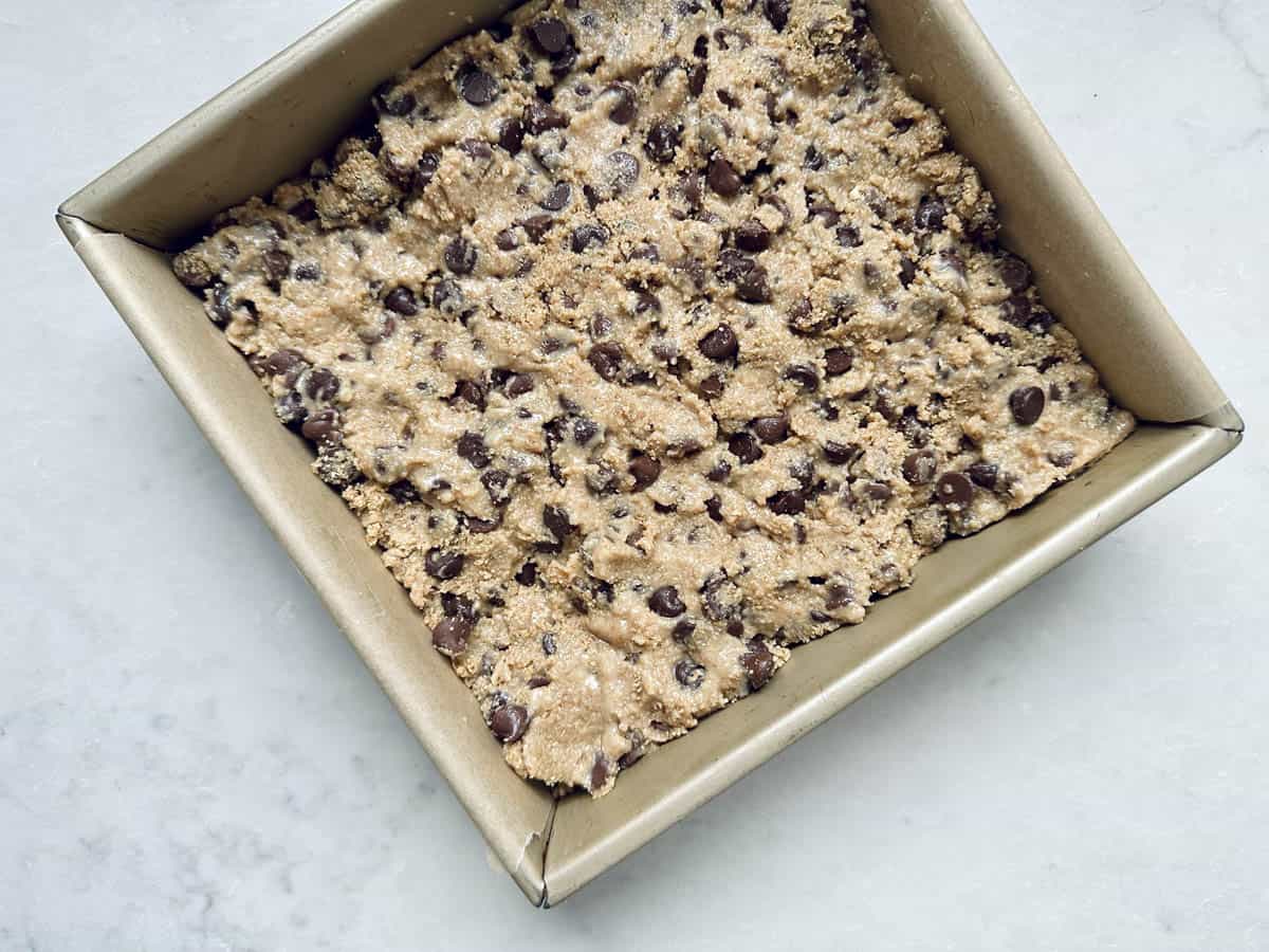 Unbaked chocolate chip cookie dough, reminiscent of the effortless 1-2-3 Snap Brownies, is pressed into a parchment-lined square baking pan, ready to go into the oven.