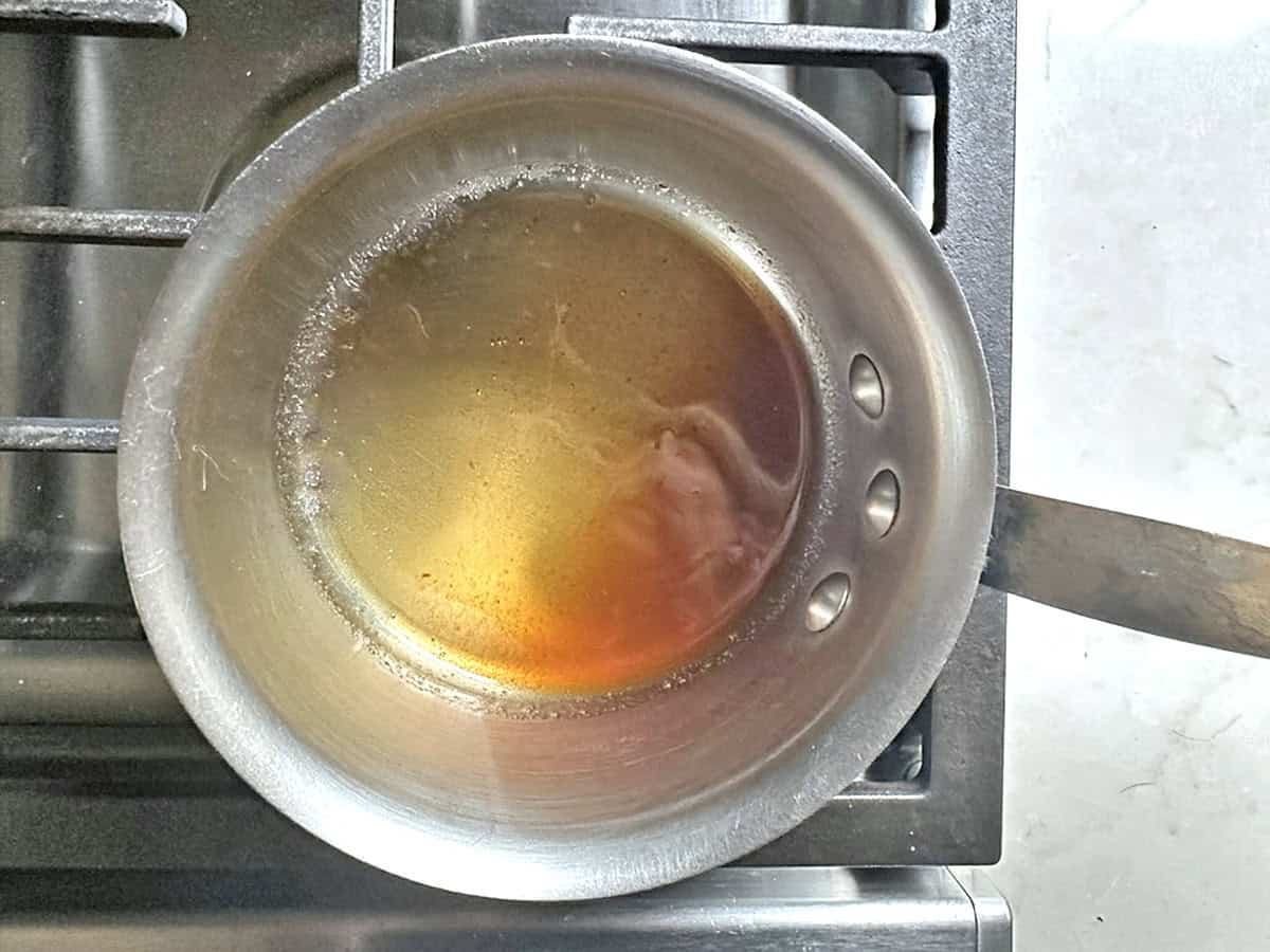 A small metal saucepan with brown liquid simmers on a stovetop, with a wooden handle extending to the right. The light reflects off the surface of the liquid, showing some bubbles around the edges, reminiscent of preparing a warm apple cider reduction for Apple Cider Snickerdoodles.