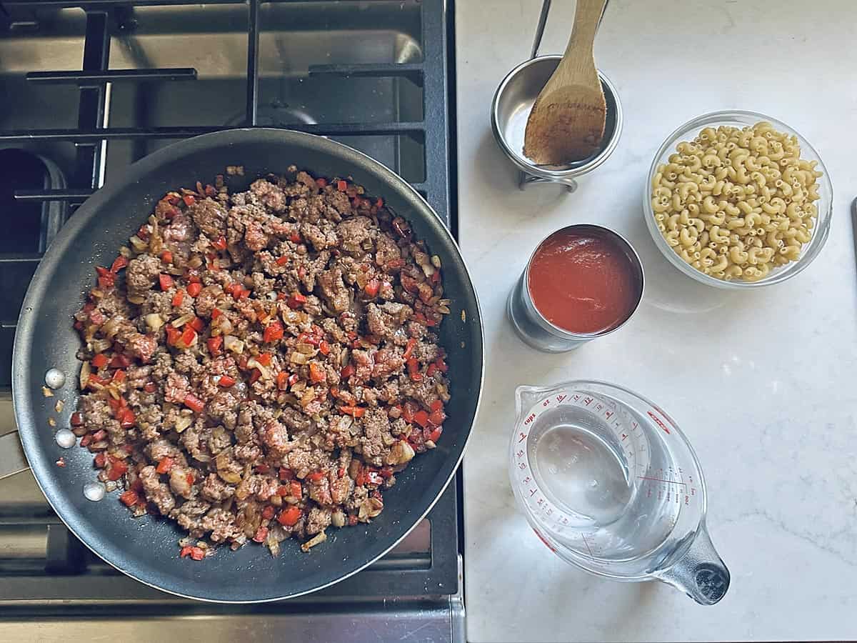 A skillet with cooked ground beef and diced veggies on a stove, next to bowls of dry macaroni, tomato sauce, and other ingredients for Skillet Cheesy Chili Mac on a white countertop.