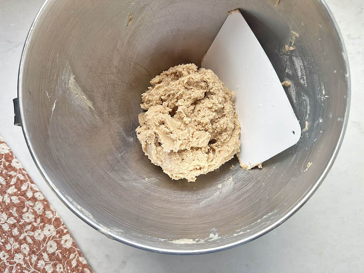 A metal mixing bowl with a ball of Apple Cider Snickerdoodles cookie dough and a white plastic dough scraper inside, placed on a light countertop next to a red patterned cloth.