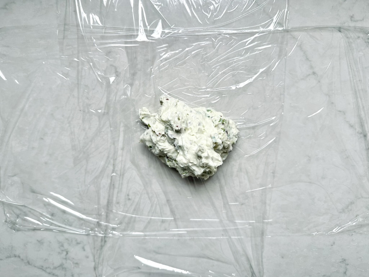 A dollop of herbed cream cheese, inspired by Vivian Howard’s Party Magnet, sits on plastic wrap atop a marble surface, ready to be wrapped.