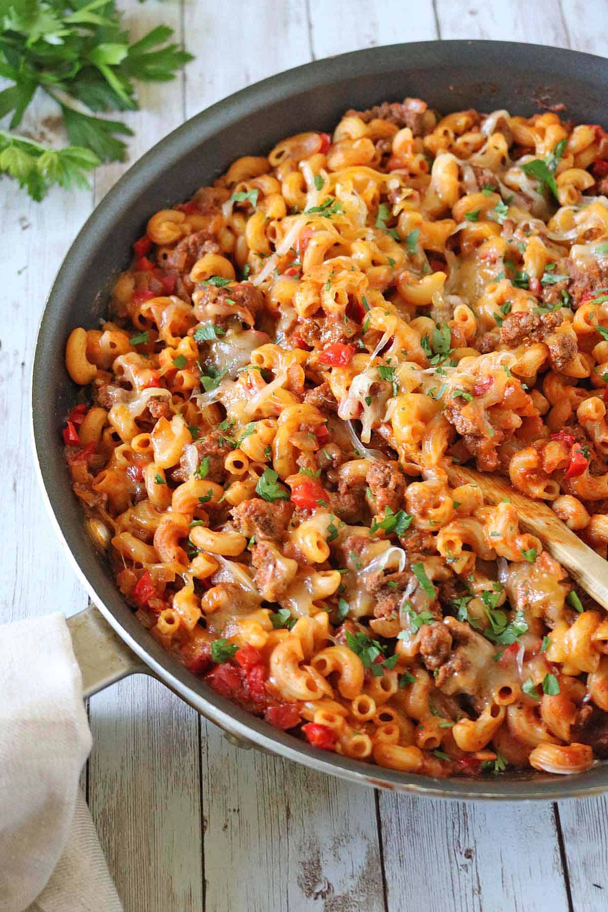 A skillet filled with Skillet Cheesy Chili Mac—macaroni pasta, ground meat, diced tomatoes, and chopped parsley—served piping hot with a wooden spoon on the side.