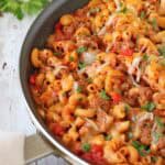 A skillet of Skillet Cheesy Chili Mac—macaroni pasta, ground beef, diced tomatoes, and melted cheese—garnished with chopped parsley sits on a rustic wooden surface.