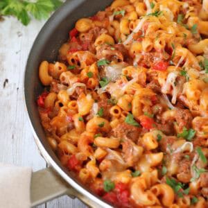 A skillet of Skillet Cheesy Chili Mac—macaroni pasta, ground beef, diced tomatoes, and melted cheese—garnished with chopped parsley sits on a rustic wooden surface.