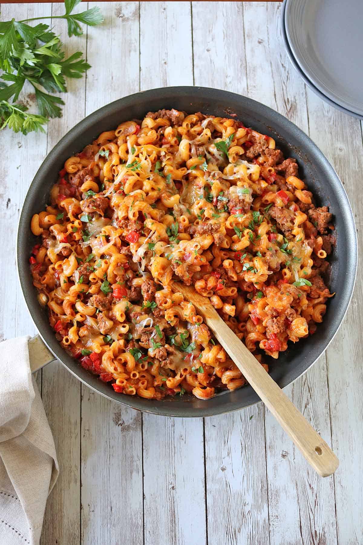 A skillet of Skillet Cheesy Chili Mac&mdash;cheesy macaroni pasta, ground meat, tomatoes, and herbs&mdash;with a wooden spoon, parsley garnish, and a napkin on a rustic wooden table.