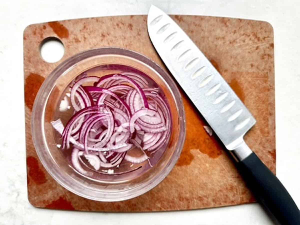 A bowl of thinly sliced red onions sits on a brown cutting board next to a sharp knife with a black handle.