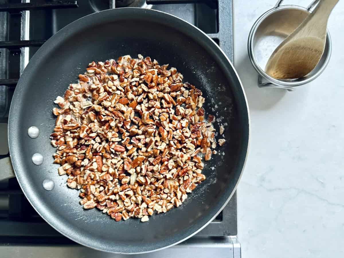 Chopped pecans toasting in a black skillet on a stove, with a small metal pitcher and a wooden spoon on a white countertop nearby—just the inviting scene you’d expect when preparing Vivian Howard’s Party Magnet.