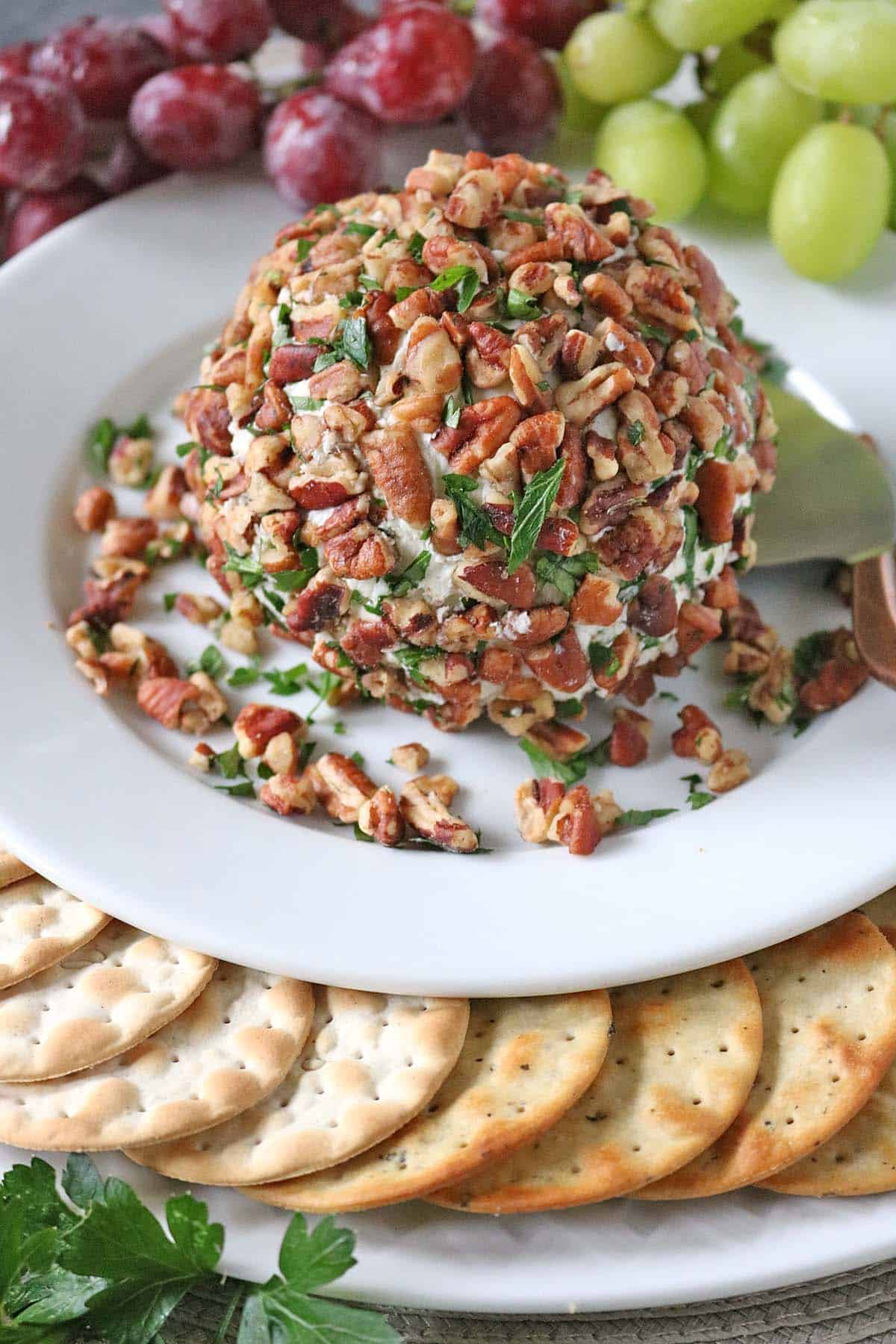 A cheese ball—Vivian Howard’s Party Magnet—coated with chopped pecans and herbs sits on a white plate, surrounded by round crackers and red and green grapes.