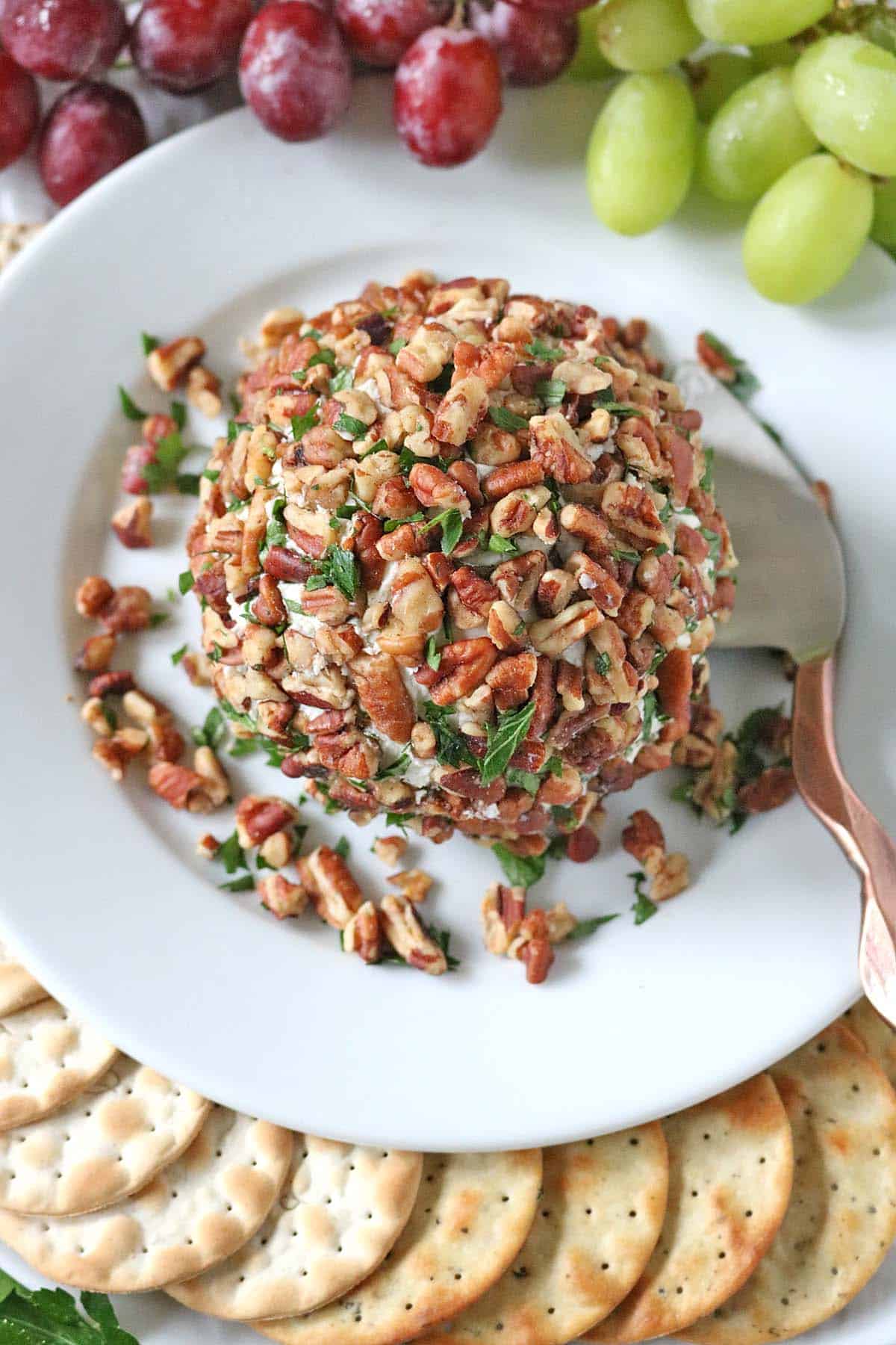 A cheese ball coated in chopped pecans and herbs, reminiscent of Vivian Howard’s Party Magnet, sits on a white plate surrounded by round crackers, red and green grapes, and a cheese knife.