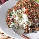 A cheese ball inspired by Vivian Howard’s Party Magnet sits on a white plate, coated with chopped pecans and herbs, and surrounded by crackers and grapes. A spoon has taken a portion out of the cheese ball, revealing its creamy interior.