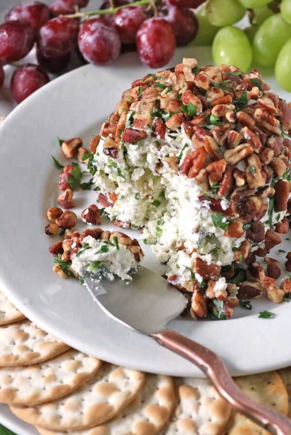 A cheese ball inspired by Vivian Howard’s Party Magnet sits on a white plate, coated with chopped pecans and herbs, and surrounded by crackers and grapes. A spoon has taken a portion out of the cheese ball, revealing its creamy interior.