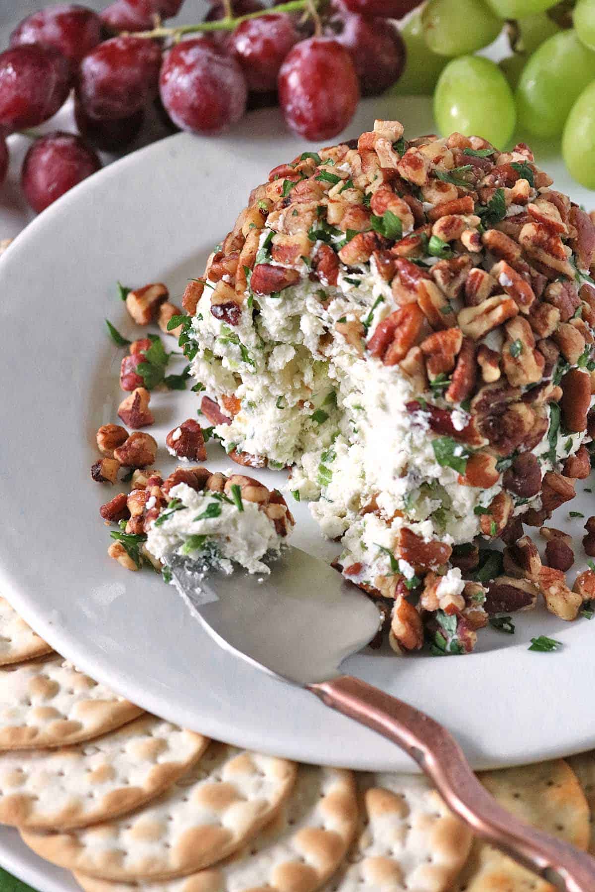 A cheese ball inspired by Vivian Howard’s Party Magnet sits on a white plate, coated with chopped pecans and herbs, and surrounded by crackers and grapes. A spoon has taken a portion out of the cheese ball, revealing its creamy interior.