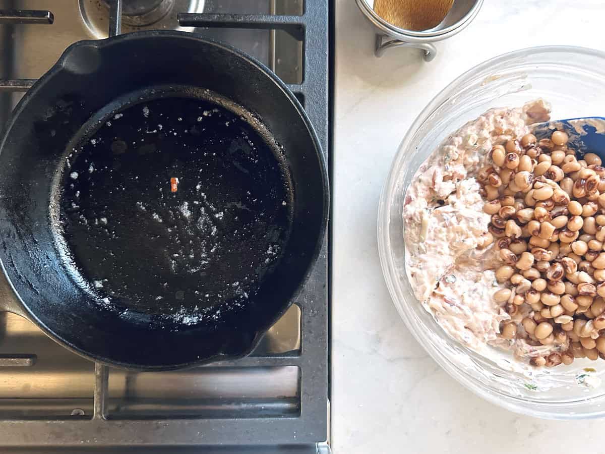 A black cast-iron skillet on a stovetop next to a glass bowl filled with a creamy baked black-eyed pea dip.