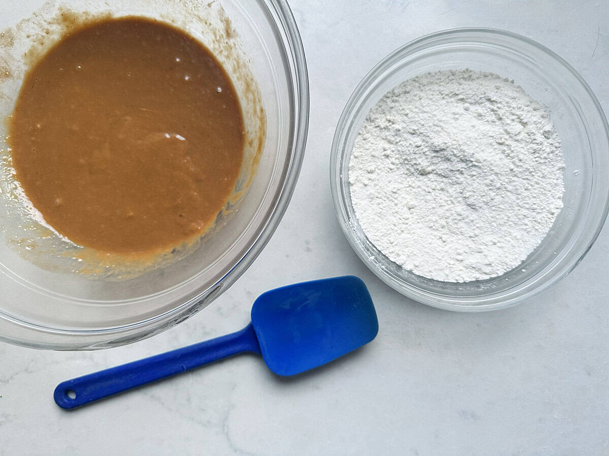 Two glass bowls on a white surface, one containing brown batter and the other containing white flour, with a blue spatula between them.