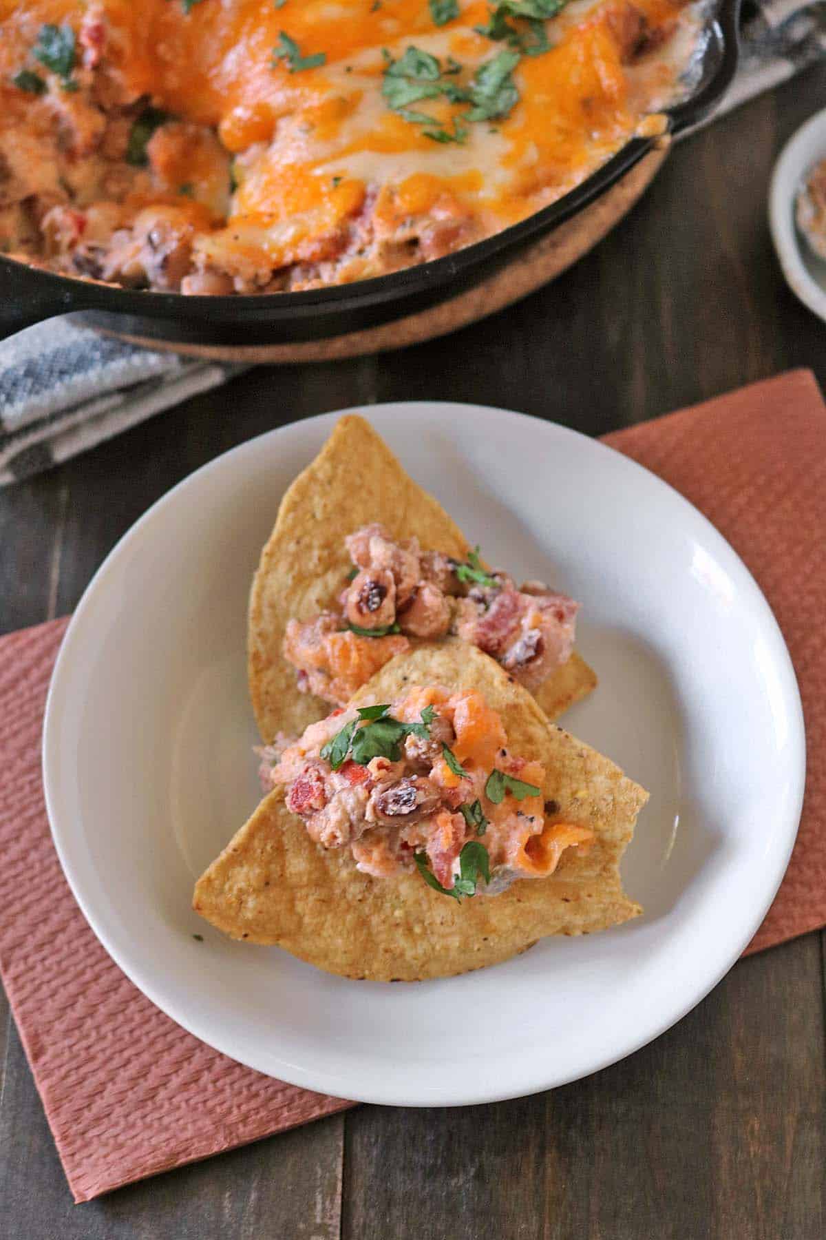 Two tortilla chips topped with a creamy baked black-eyed pea dip on a white plate, with a cast-iron skillet of the same dip in the background.