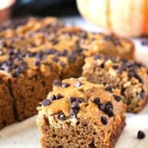 A square piece of pumpkin chocolate chip bread in the foreground, with more slices and a whole pumpkin in the background.