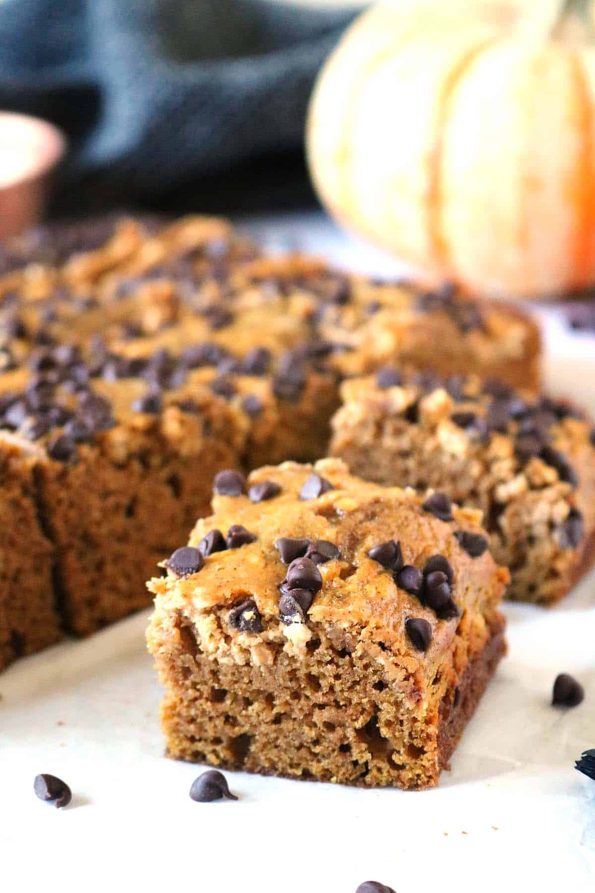 A square piece of pumpkin chocolate chip bread in the foreground, with more slices and a whole pumpkin in the background.