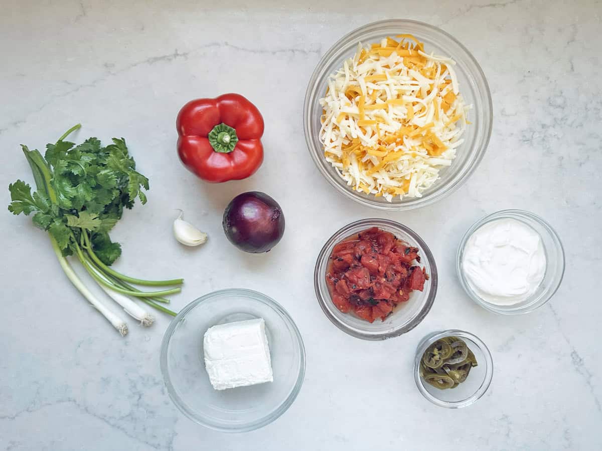 Ingredients for a baked black-eyed pea dip arranged on a counter, including shredded cheese, diced tomatoes, sour cream, jalapeño slices, a red bell pepper, red onion, a garlic clove, cream cheese, and fresh cilantro.